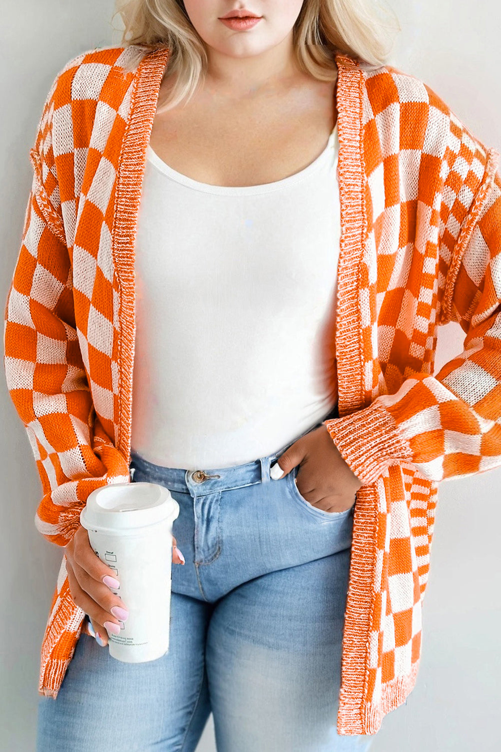 Person wearing an orange and white checkered cardigan over a white top and blue jeans, holding a white cup.