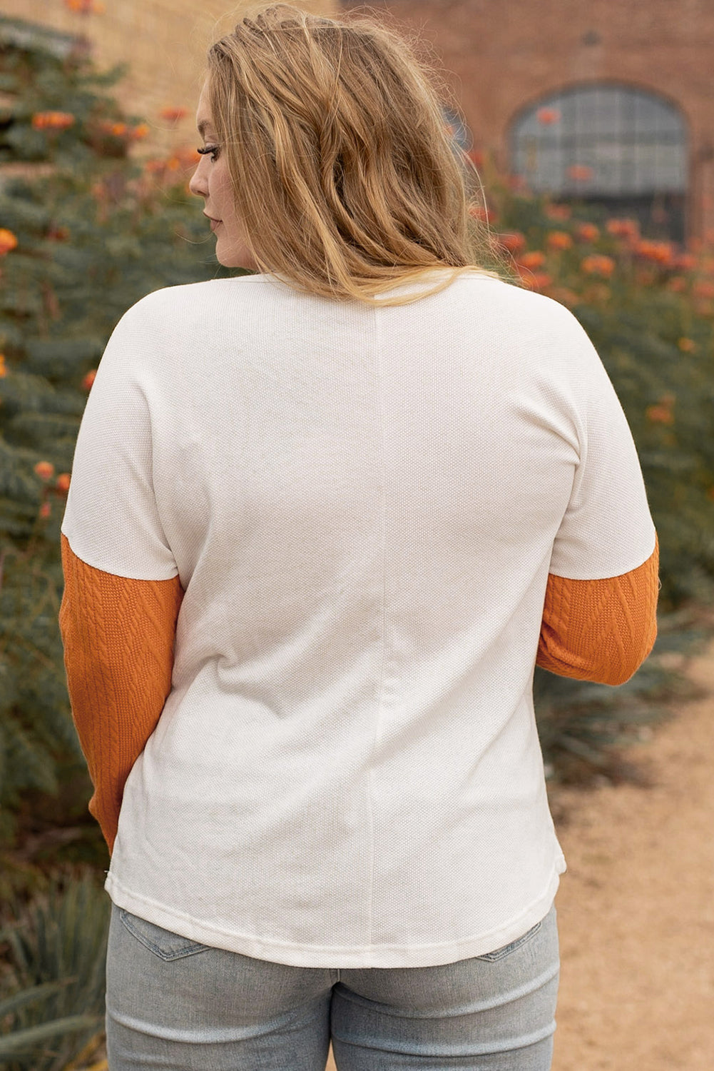 Person wearing a white shirt with orange sleeves in an outdoor setting with trees and a building.
