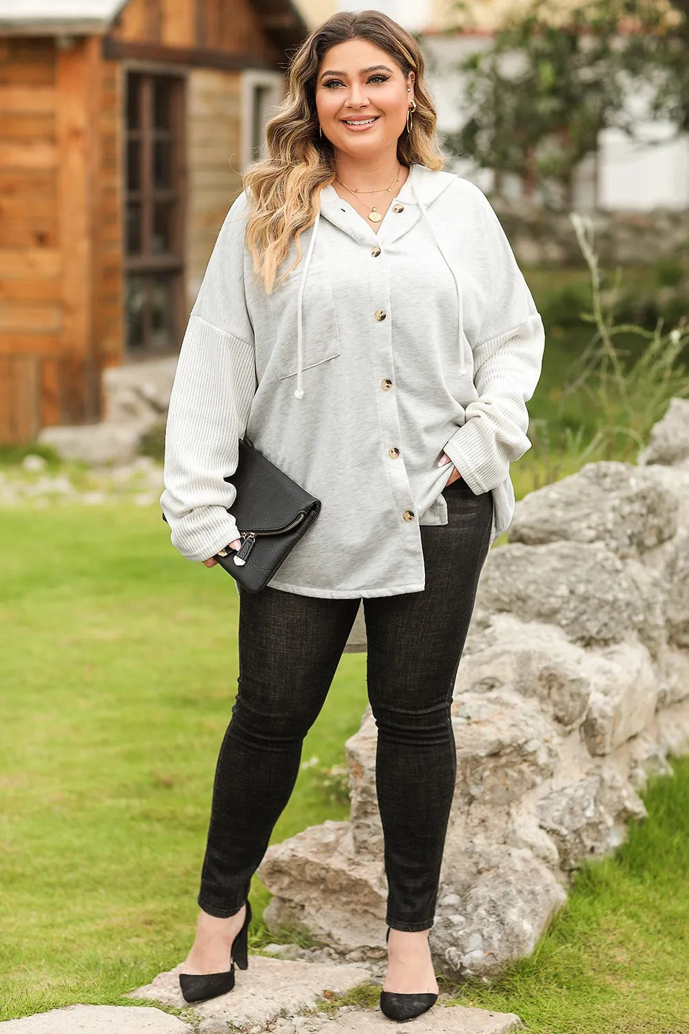 Woman wearing a light gray hoodie and black pants standing outdoors near a wooden cabin.