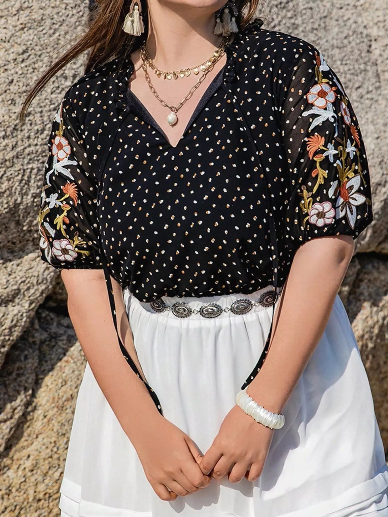 Woman wearing a black polka dot blouse with floral sleeves against a stone wall.