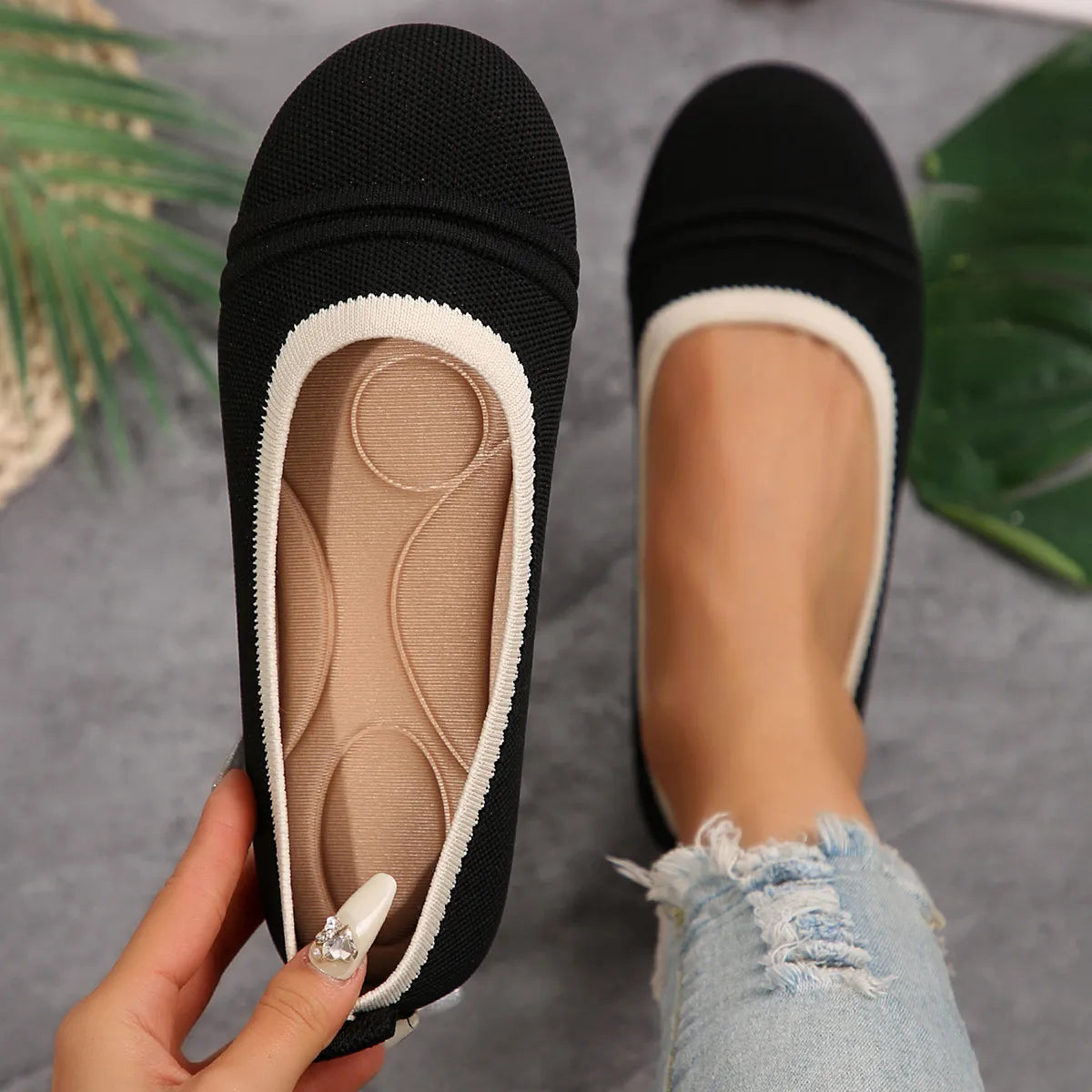 Black and white flat shoes with a close-up of the sole, held by a hand against a gray background with green leaves.
